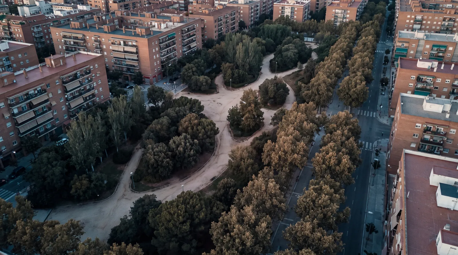 Parque urbano con arboleda junto a bloques de viviendas de ladrillo y una avenida, visto desde el aire a primera hora.