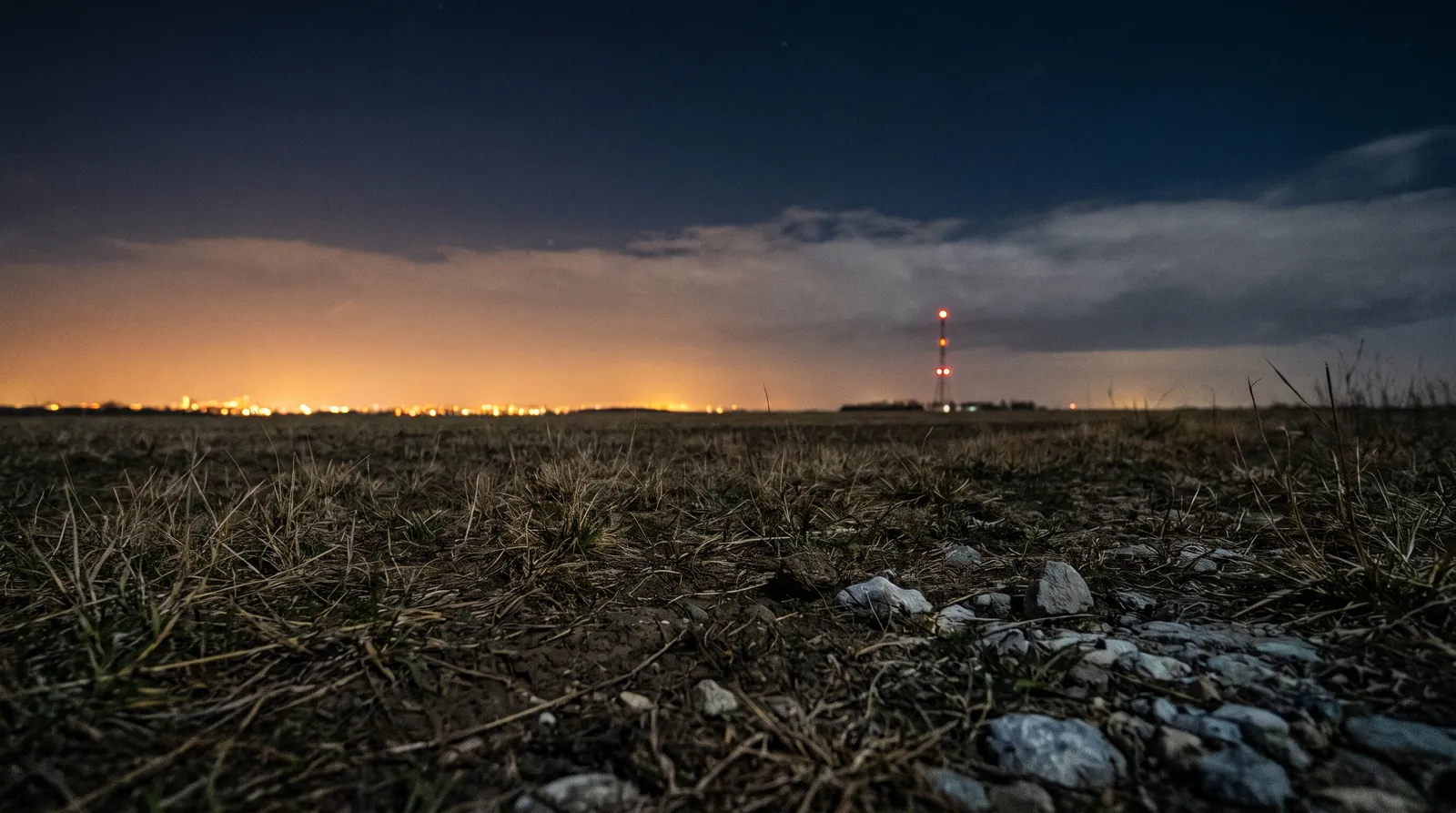 Campo abierto de noche con el resplandor de una ciudad en el horizonte y las luces rojas de una antena lejana.