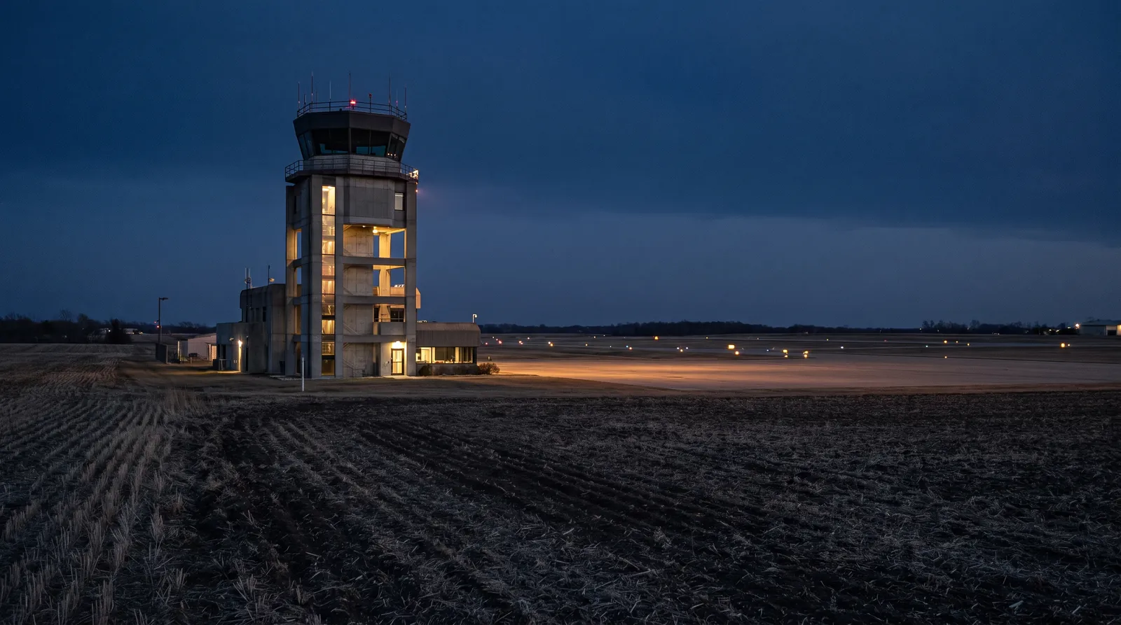 Torre de control de un aeropuerto regional al anochecer, con las luces de pista encendidas, vista desde un campo cercano.