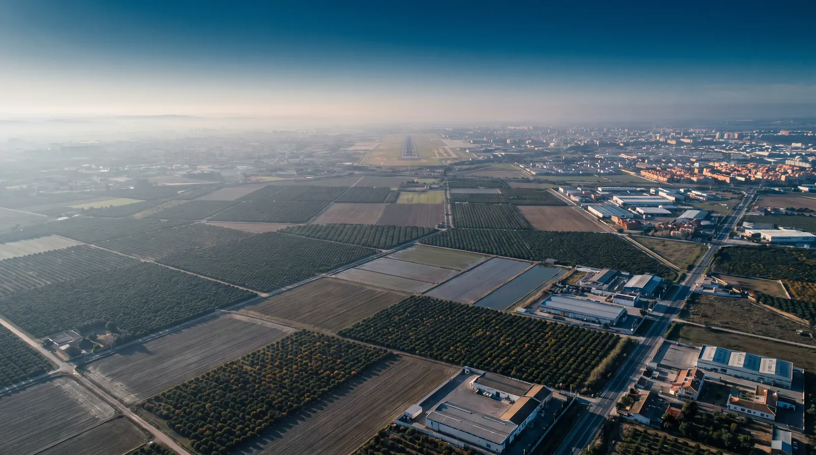 Vega de naranjales y parcelas de regadío con la pista de un aeropuerto a lo lejos, vista desde el aire.